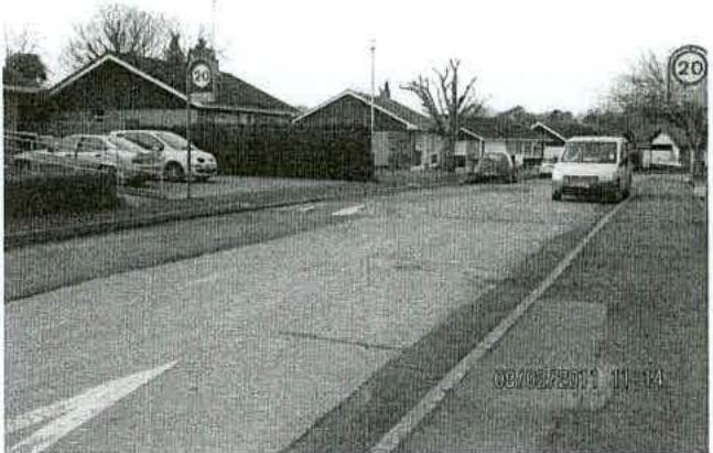 A black and white photograph showing a street scene with a commercial building, parked cars, and a white van driving on the road.