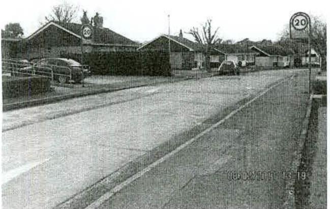 A grainy black and white street-level photograph showing a road with parked cars and single-story commercial buildings in the background.