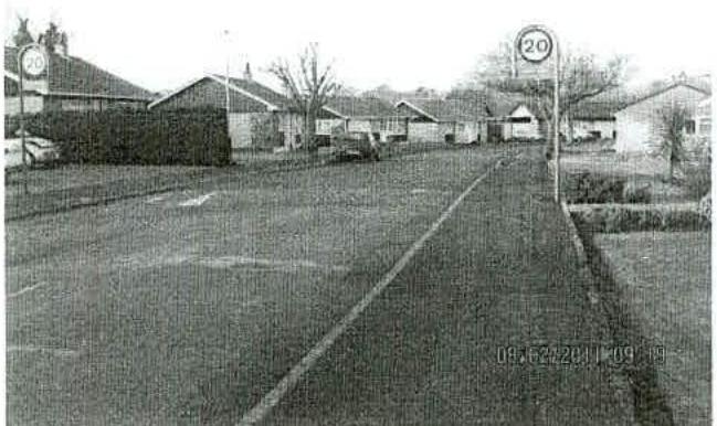 A grainy black and white photograph showing a street scene with houses, parked cars, and 20 mph speed limit signs.