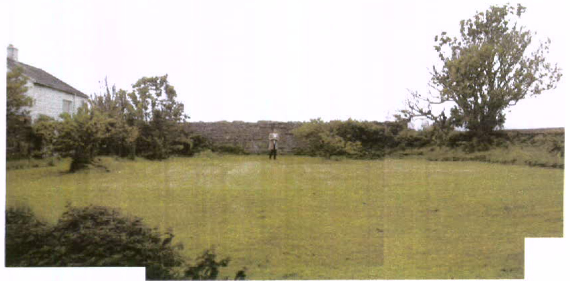 A photograph showing a grassy field or yard enclosed by a stone wall, with a white building visible on the left and trees in the background.