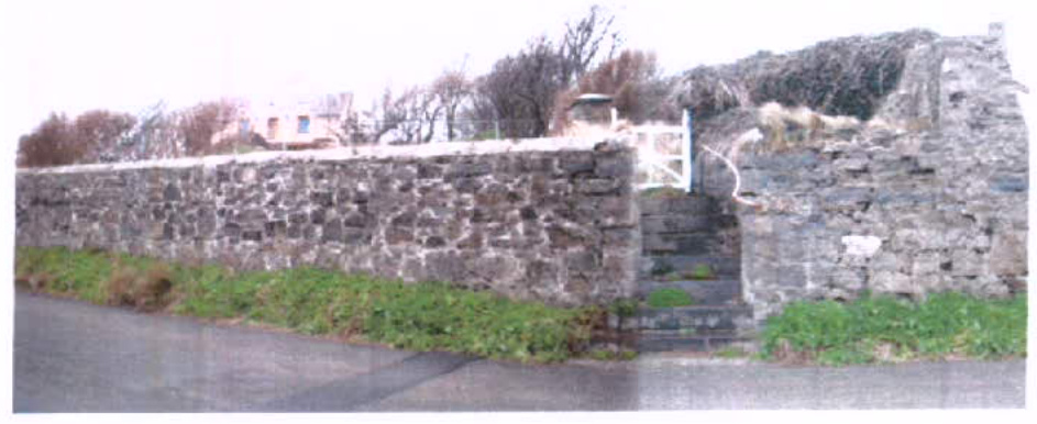 A photograph showing a long, rough stone boundary wall running along a roadside, featuring a white gate and stone steps leading up to a property.