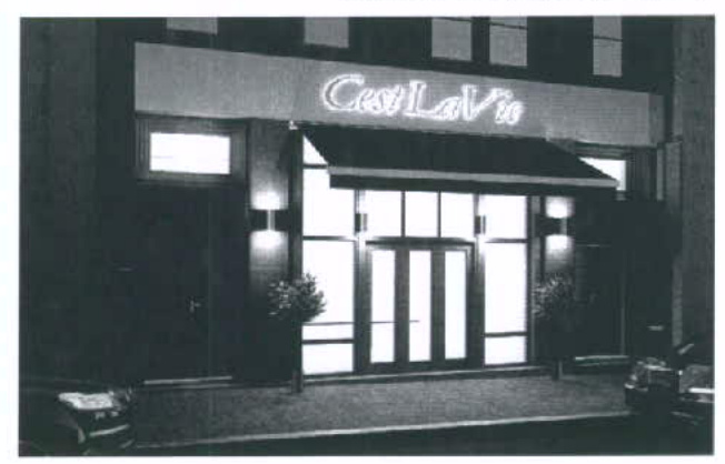 A black and white photograph showing the illuminated night-time exterior of a commercial establishment with a sign reading 'C'est La Vie' and a canopy over the entrance.