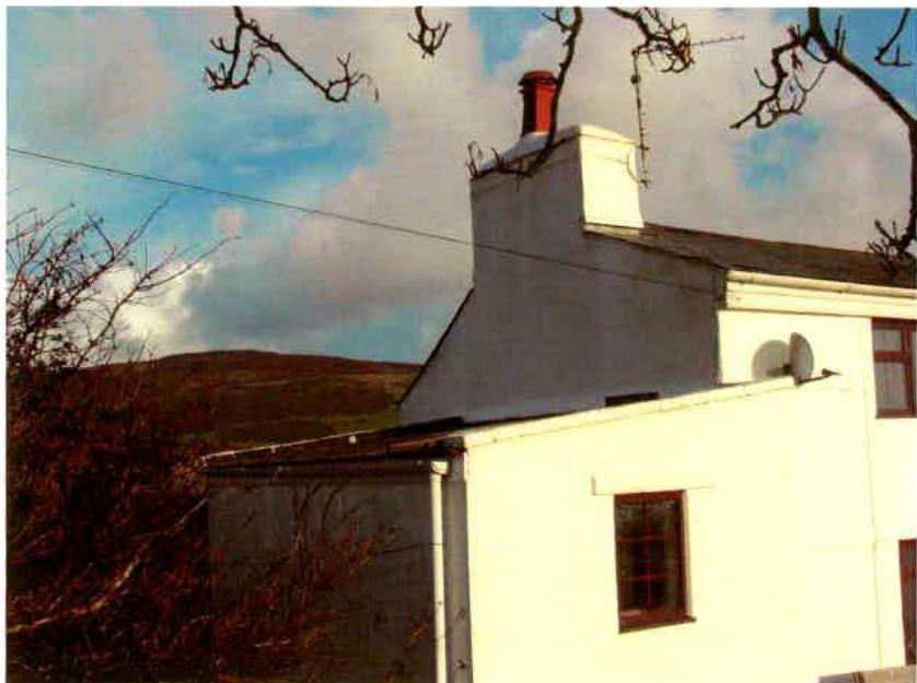 A photograph showing the side elevation of a white detached house with a chimney and satellite dish, set against a rural hillside background.