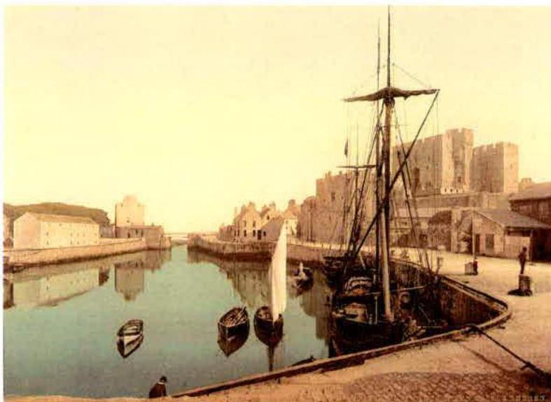 A historical sepia-toned photograph depicting a harbor scene with a large sailing ship, smaller boats, and stone buildings including a castle in the background.