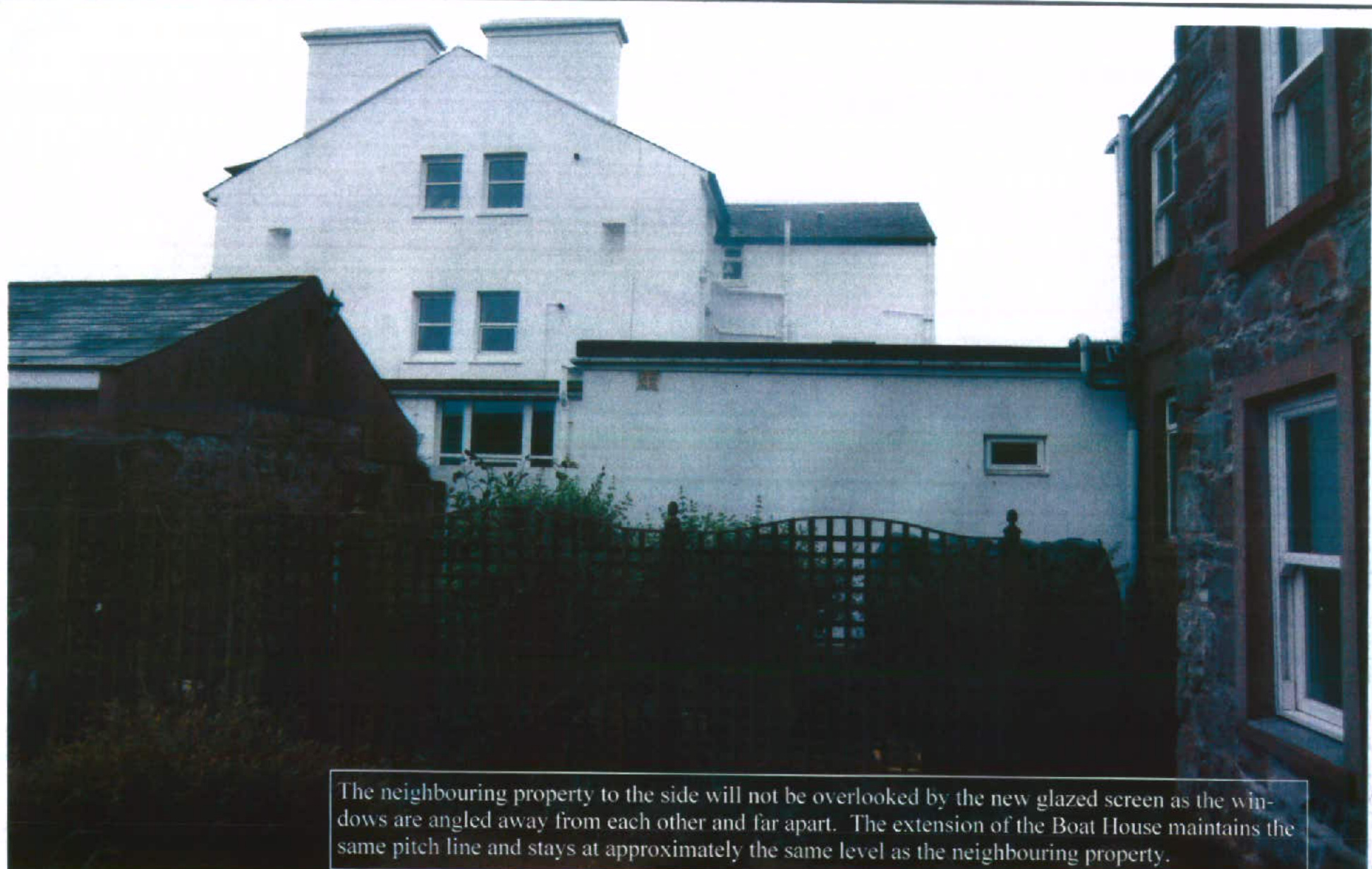 A photograph showing a white two-storey house and a lower outbuilding structure, with a stone neighboring property visible on the right.