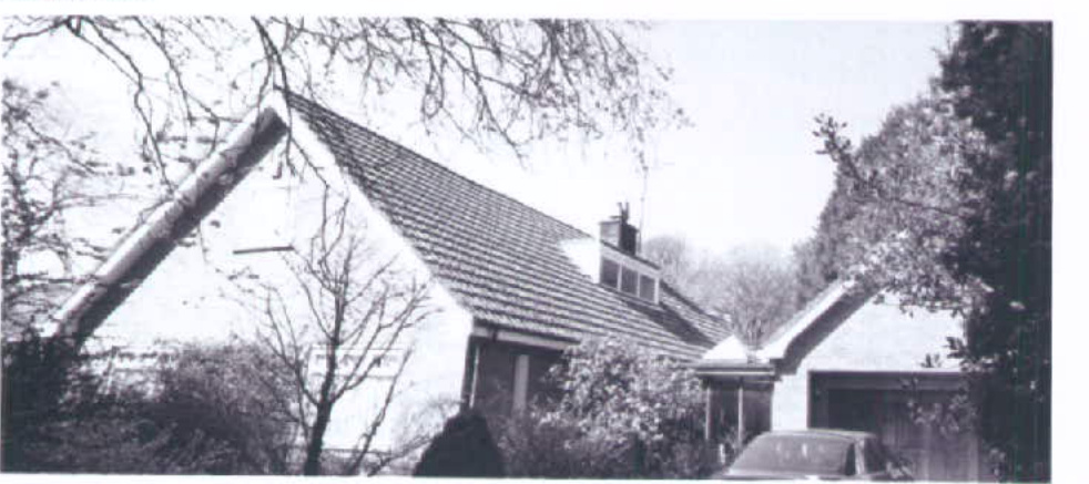 A black and white photograph showing a detached bungalow with a pitched roof and an attached garage, surrounded by trees and vegetation.