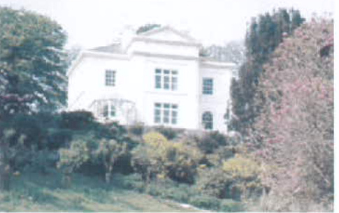 A grainy photograph showing a large white two-story detached house situated on a slope surrounded by trees and vegetation.