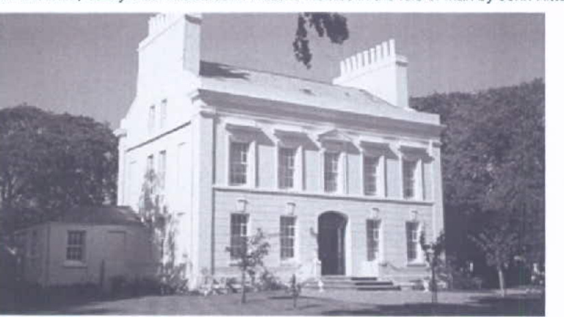 A black and white photograph showing a large, two-story detached house with a symmetrical facade and prominent chimneys, alongside a smaller outbuilding to the left.