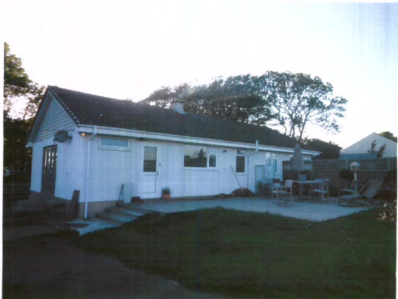 A photograph showing a white, single-story bungalow with a tiled roof, featuring a rear patio area and lawn.