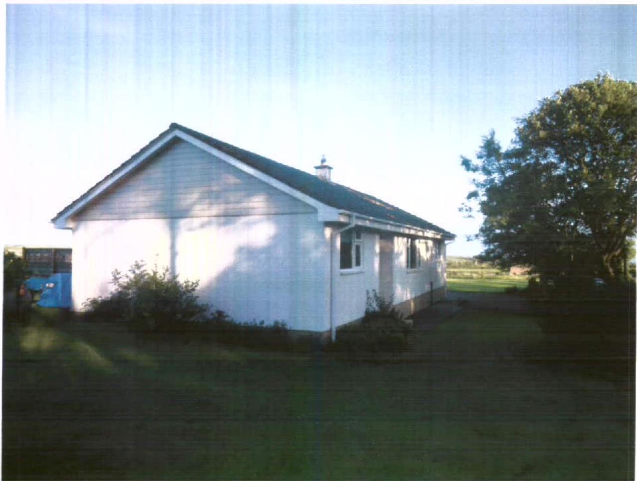 A photograph of a white, single-story detached bungalow with a pitched roof situated in a grassy area with trees.