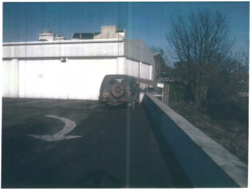 A photograph showing the rear exterior of a white commercial building with a vehicle parked in a paved area featuring a directional arrow.