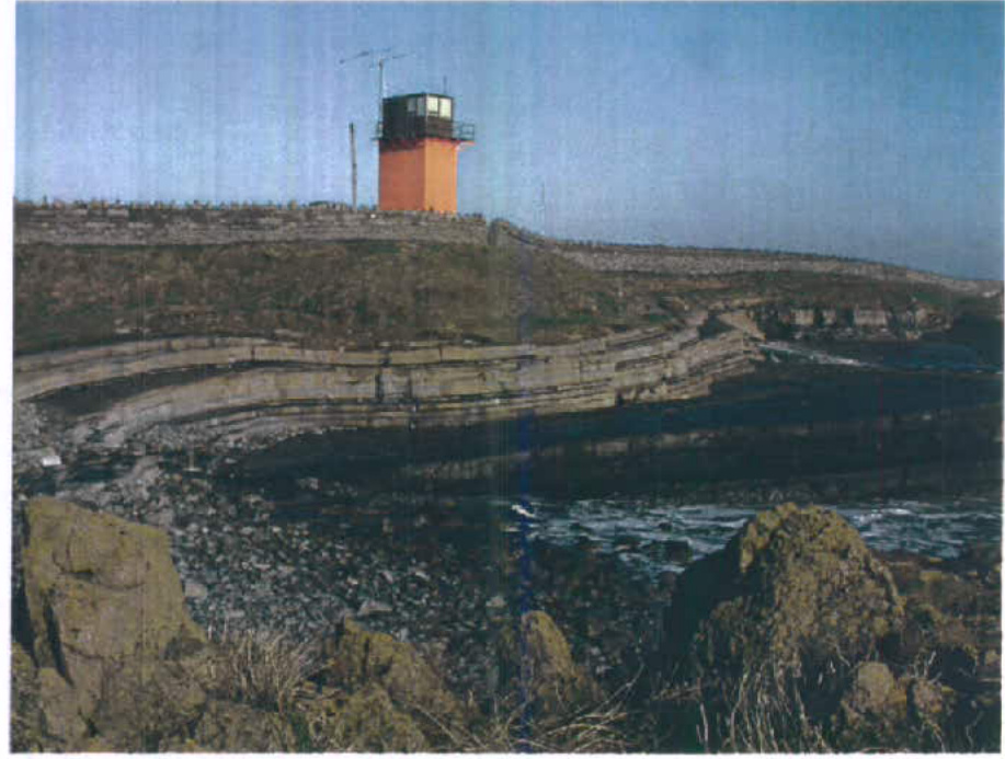 A photograph of a rocky coastal landscape featuring tiered rock formations or sea defenses leading up to a stone wall and a small orange tower structure.