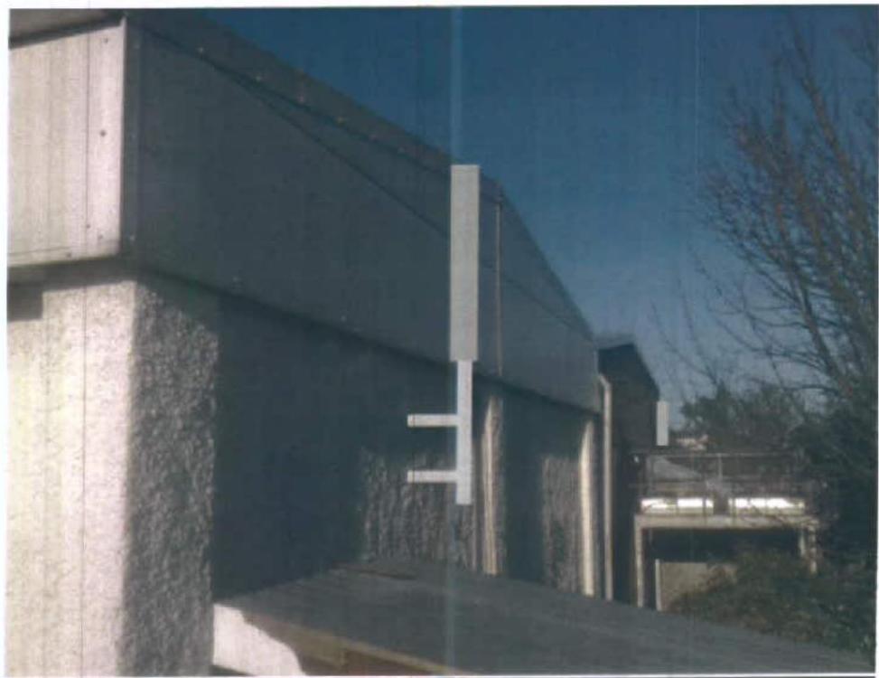 A photograph showing the side elevation of a large building with textured rendering and a flat roof overhang, set against a blue sky with trees in the background.