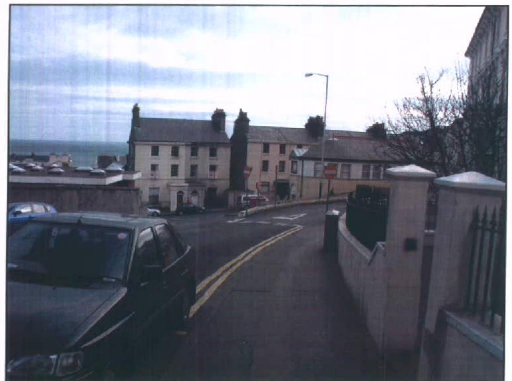 A street-level photograph showing a curved road with terraced buildings in the background and the sea visible in the distance.