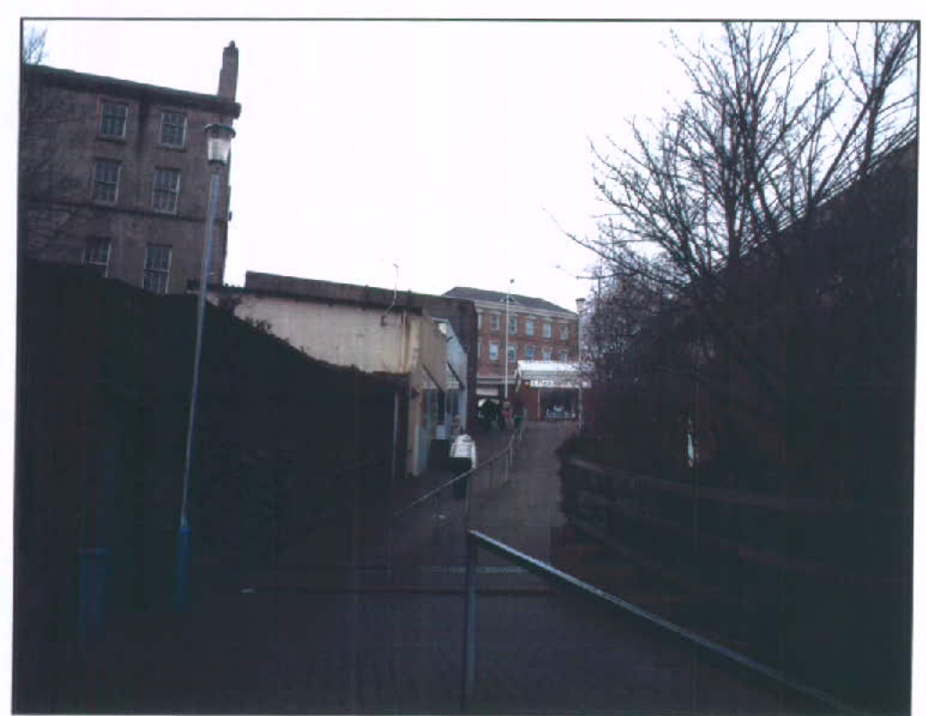 A photograph showing the existing street scene with multi-story buildings on the left and a paved path leading uphill, documenting the current site conditions.