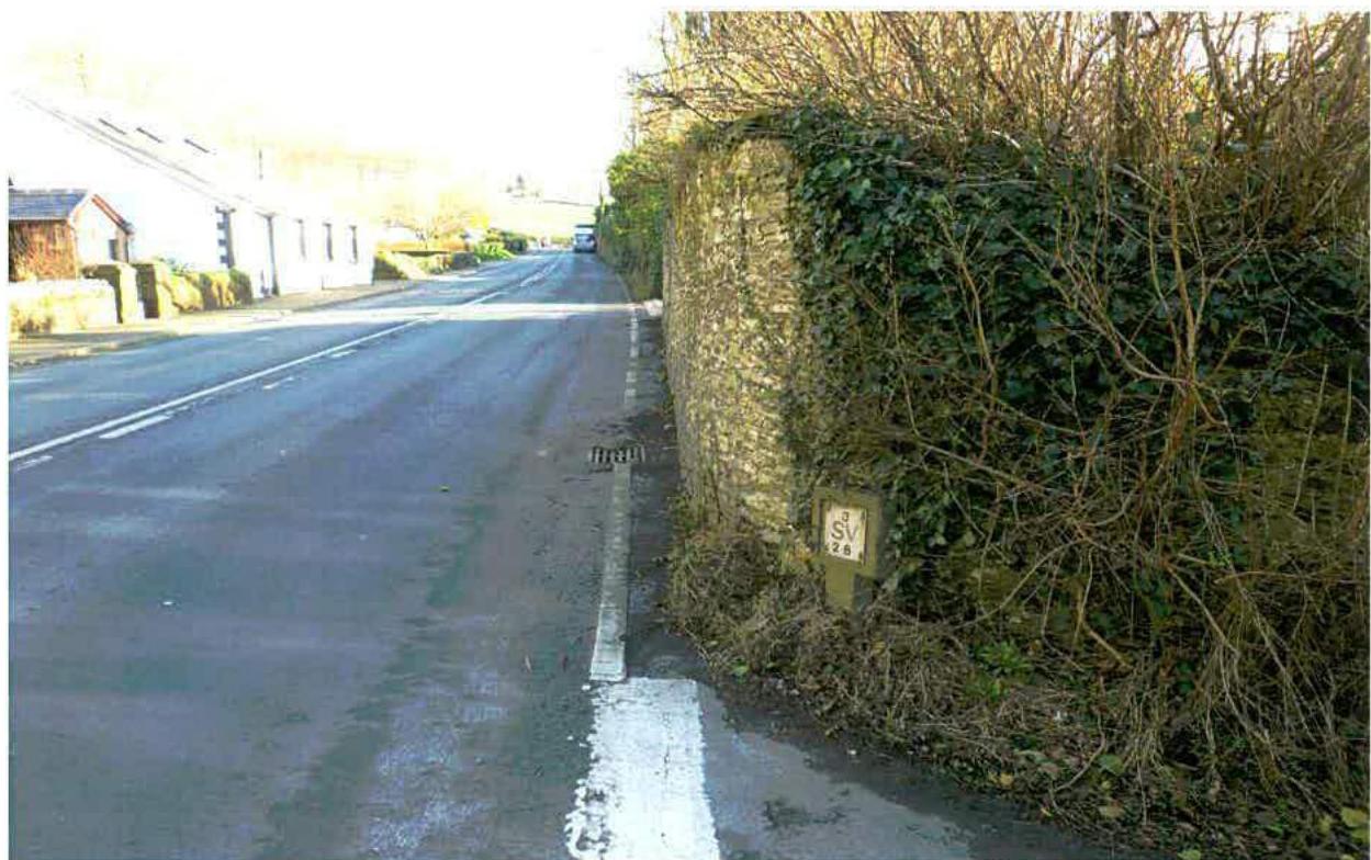 A photograph of a paved road with white markings, bordered by a stone wall covered in ivy on the right and a white building on the left.