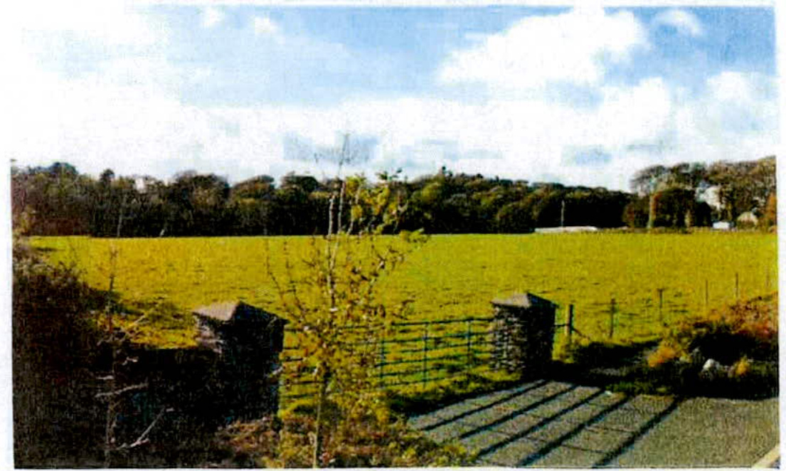 A photograph of a rural landscape featuring a large green field, stone gate pillars, and a metal gate in the foreground.