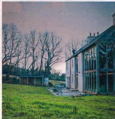 A photograph showing a detached building with a large glass extension and a smaller wooden outbuilding in the background, set against a backdrop of bare trees and a grassy lawn.