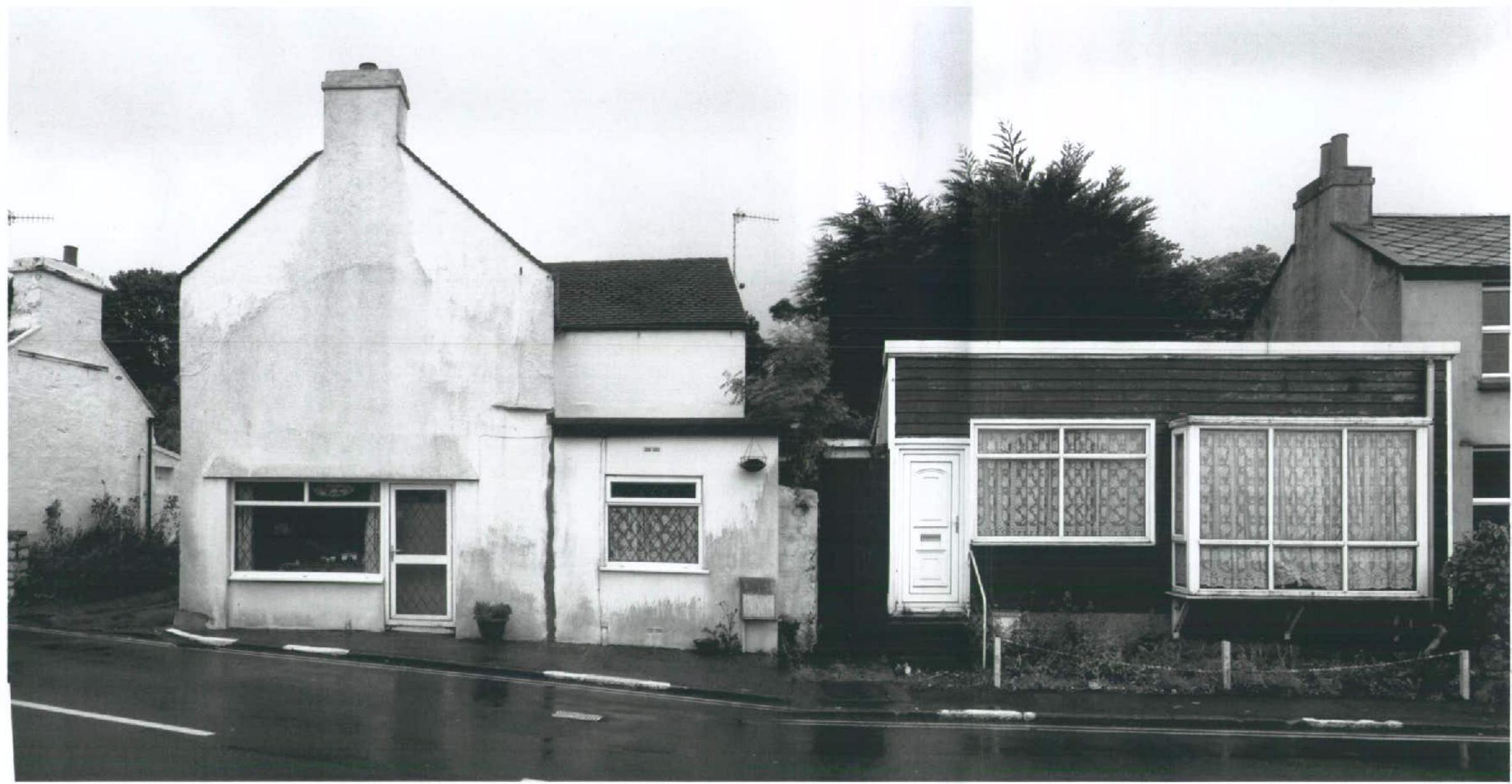 A black and white photograph showing the exterior of two adjacent buildings, including a white gabled structure and a single-story bungalow with a flat-roofed extension.