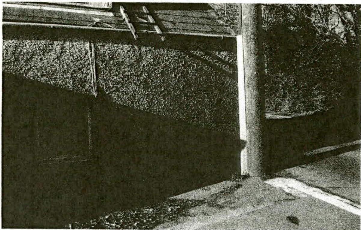 A grainy black and white photograph showing the textured exterior wall of a building with a downpipe and a paved area in the foreground.