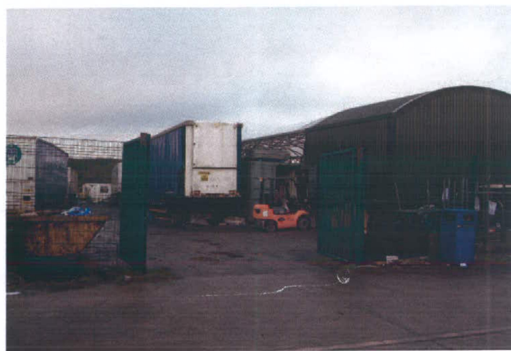 A grainy photograph showing an industrial yard with large metal sheds, shipping containers, and a forklift truck near a green gate.
