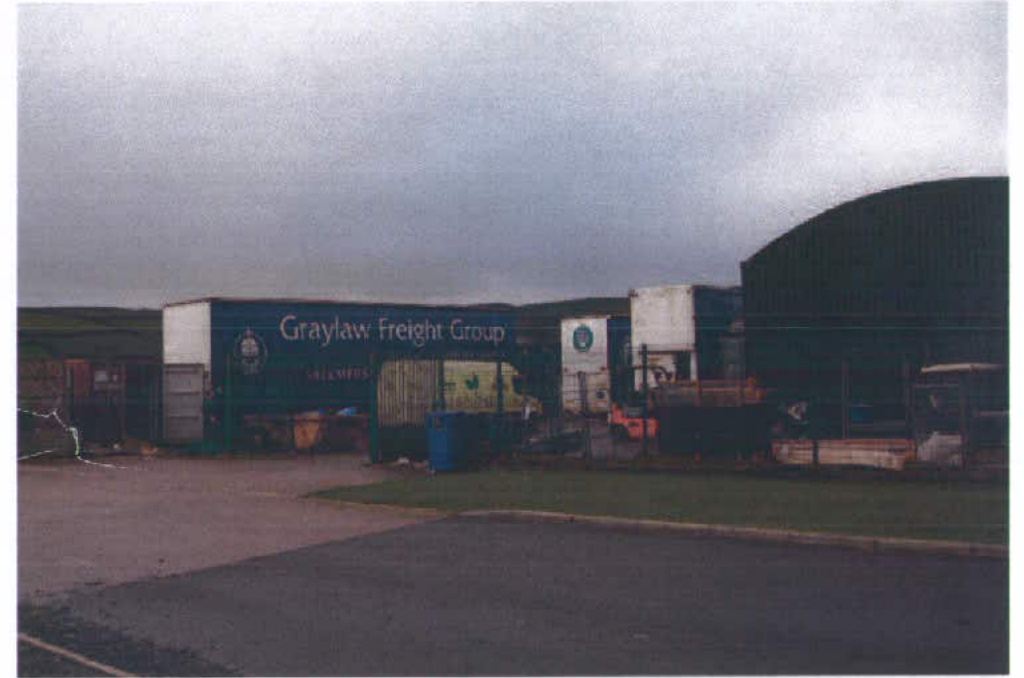 A photograph of a commercial freight depot featuring shipping containers and a large arched warehouse building under an overcast sky.