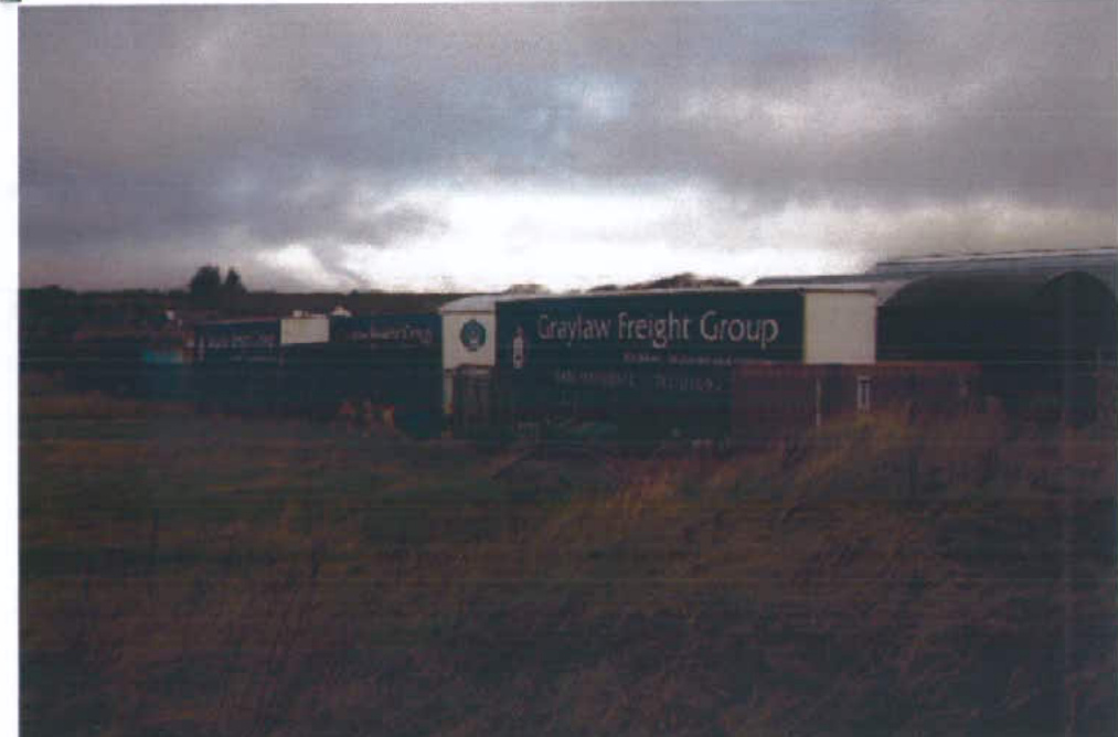 A photograph showing a commercial site with a large 'Graylaw Freight Group' sign and warehouse-style buildings in the background, viewed from a grassy foreground.