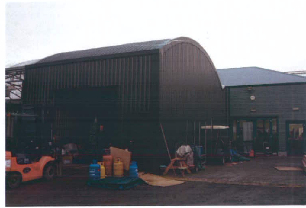 A photograph showing a large, dark, curved-roof industrial or agricultural building (resembling a Quonset hut) adjacent to a rectangular office-style structure with a forklift in the foreground.