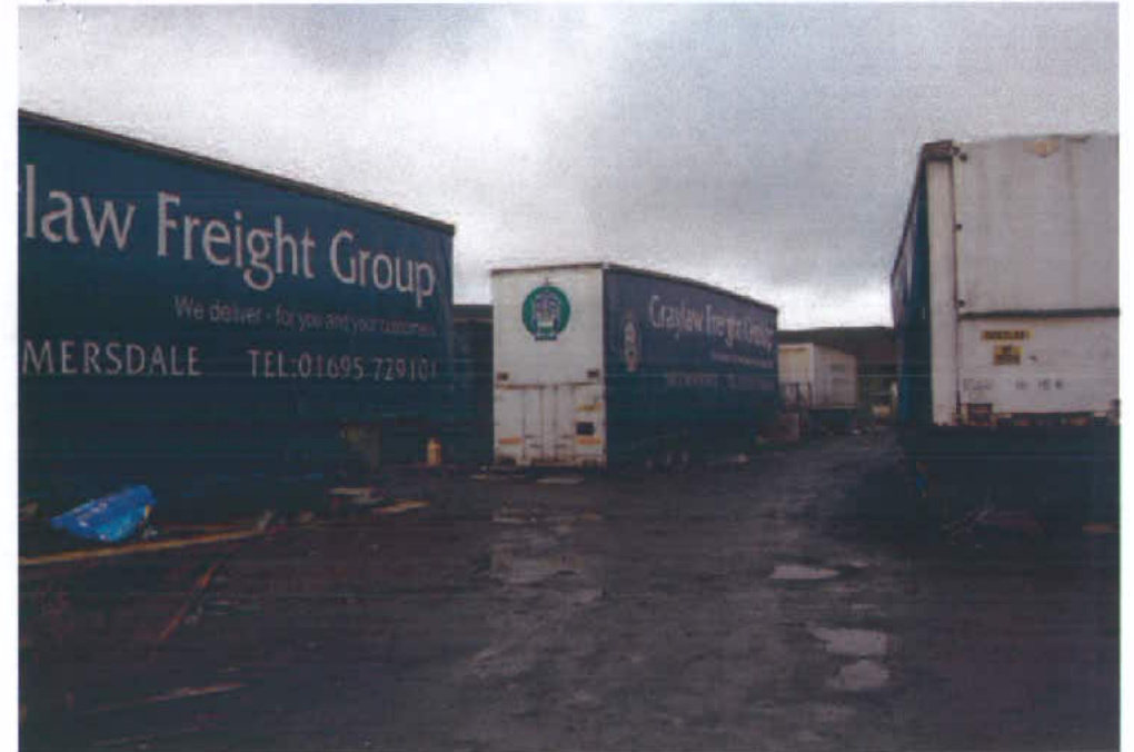 A photograph showing a commercial freight yard with large shipping containers and trailers parked on a wet paved surface.
