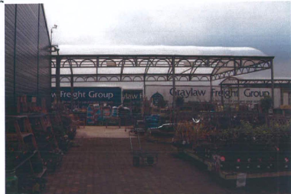 A photograph of an outdoor commercial site featuring a large metal canopy structure with a translucent roof. Signs for Graylaw Freight Group are visible in the background, along with plants and equipment in the foregr...