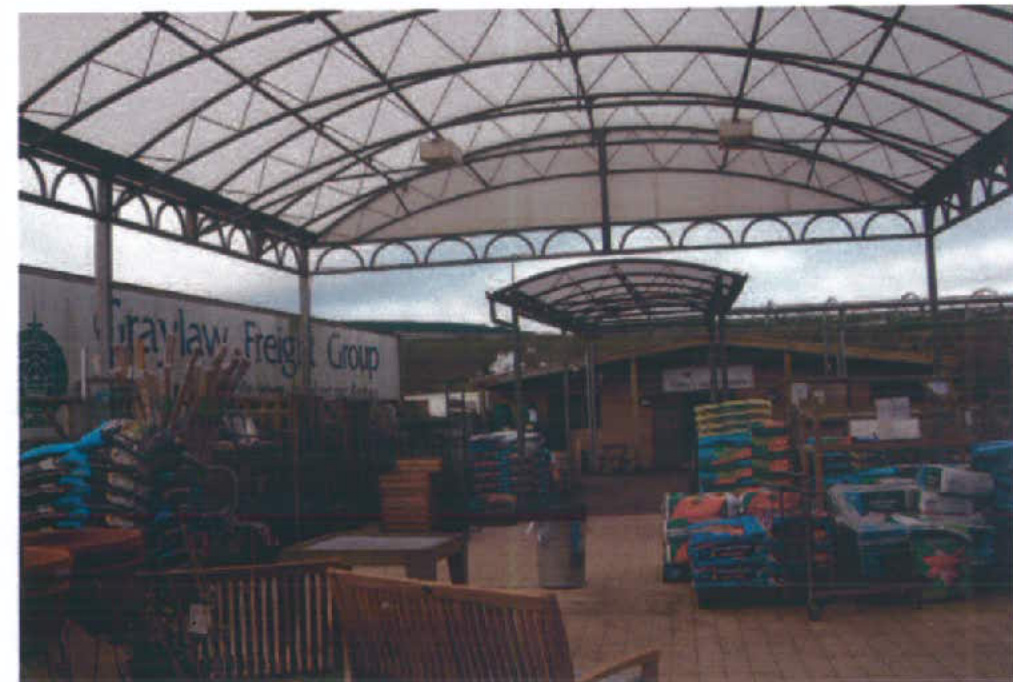 A photograph showing a large metal-framed canopy structure covering a yard area with stacked bags and pallets.