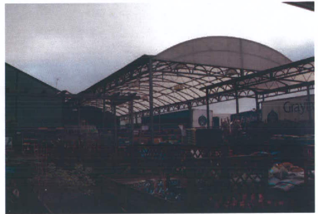 A grainy photograph showing a large steel-framed agricultural or industrial shed with a curved roof structure.