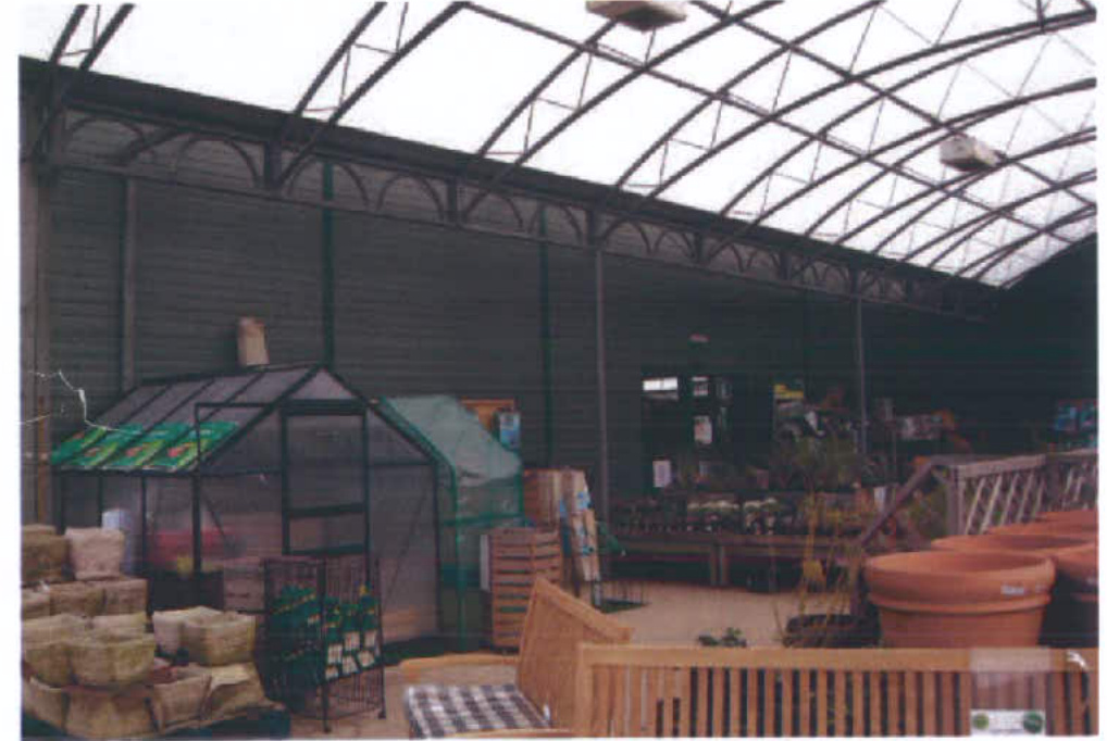 Interior photograph of a large garden center or nursery building featuring terracotta pots, stacked soil bags, and a small greenhouse structure under a high arched roof.