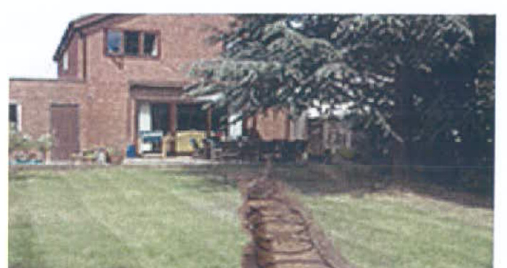 A grainy photograph showing a two-story brick house with a single-story extension, partially obscured by a large tree and a lawn in the foreground.