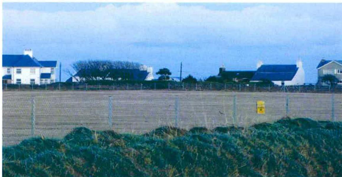 A photograph showing a large open field or sports pitch in the foreground with a fence, and existing residential houses visible in the background under a cloudy sky.