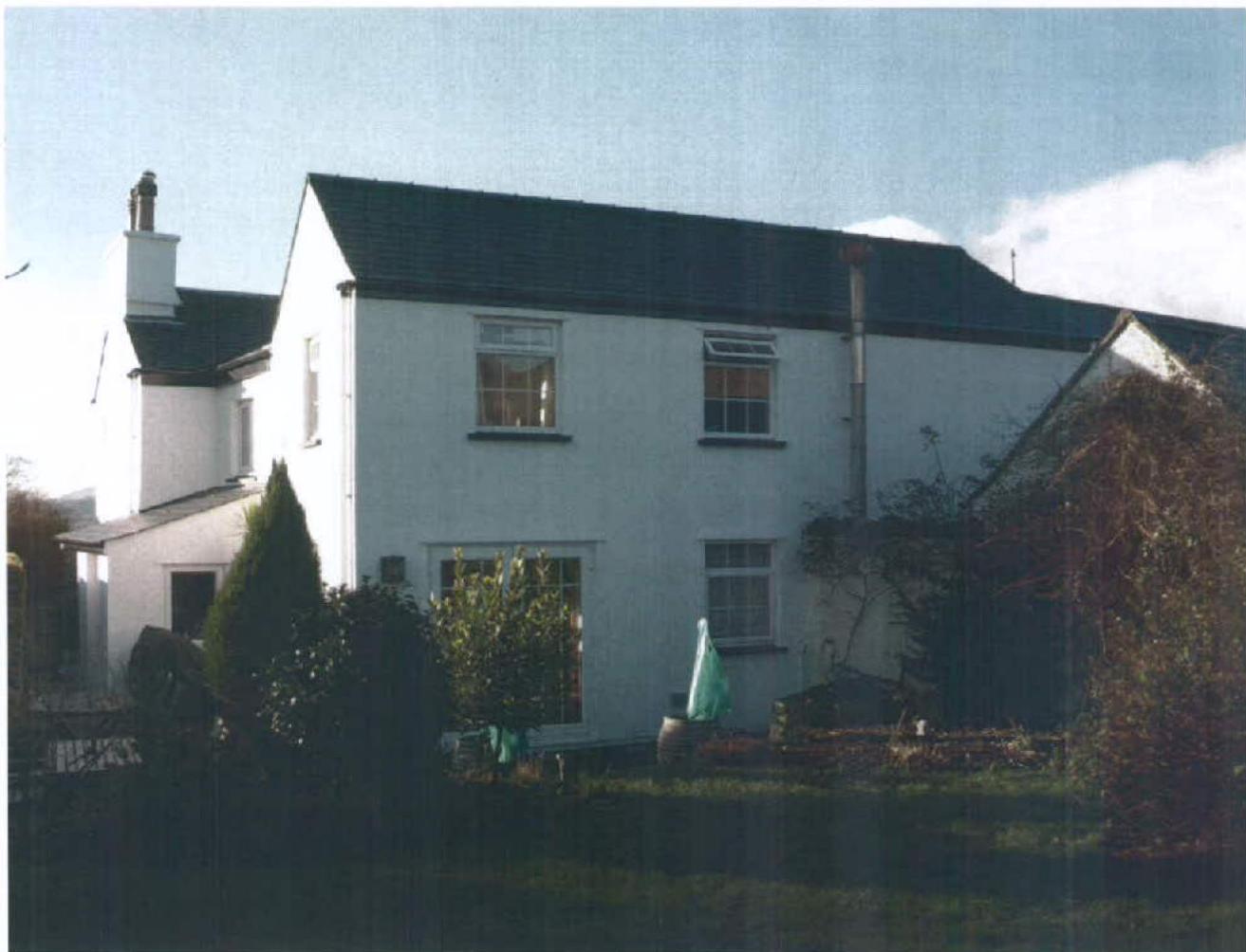 A grainy photograph showing the front exterior of a white, two-story residential house with a dark roof and a garden in the foreground.