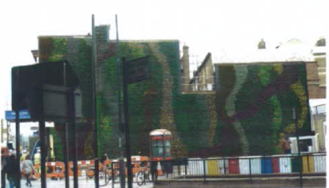 A street-level photograph showing a building facade covered in camouflage netting, with a red telephone box and construction barriers in the foreground.