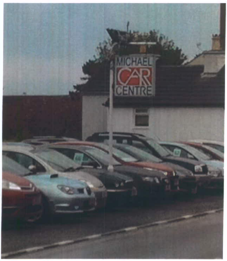A photograph showing a car dealership lot with a row of parked cars and a sign reading 'Michael Car Centre' in front of a building.
