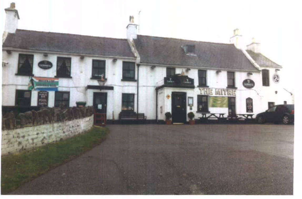 A street-level photograph of a white two-story building identified as 'The Mitre' pub, featuring multiple advertising signs on the facade and a car parked in the foreground.