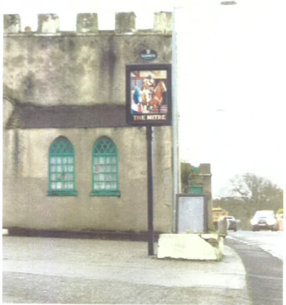 A photograph showing the exterior of a stone building, likely a pub named 'The Mitre', featuring a hanging advertising sign and arched windows.