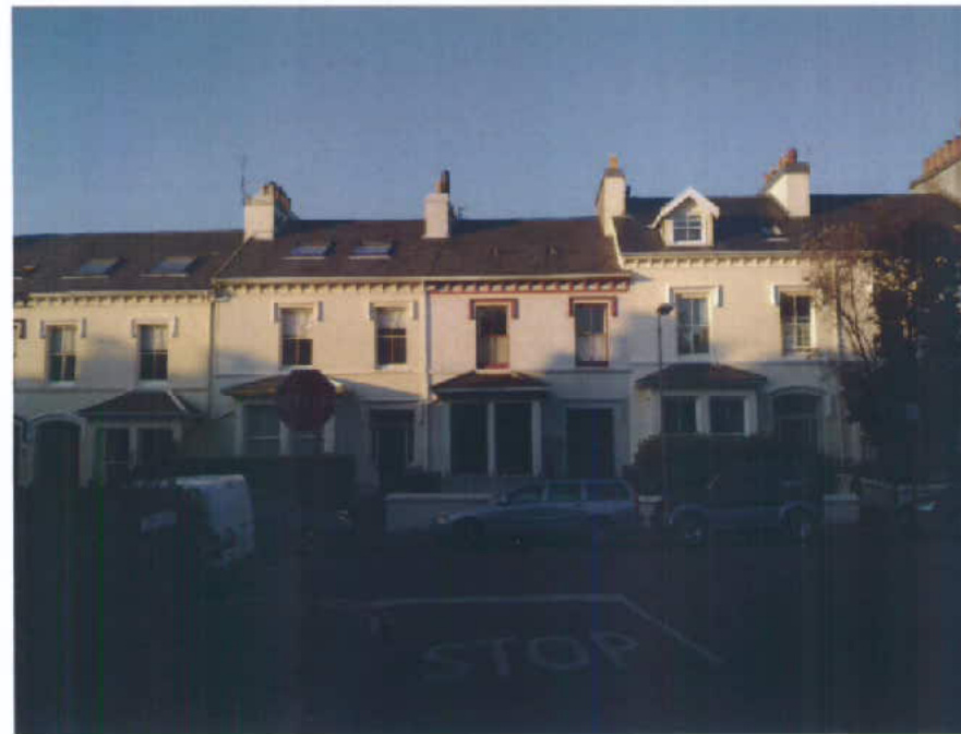 A street-level photograph showing a row of white terraced houses with dark tiled roofs and dormer windows, with cars parked in front.