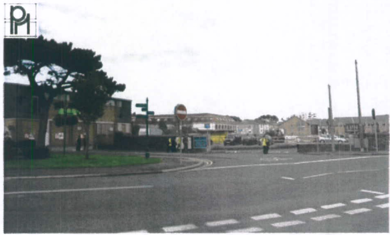 A street-level photograph showing a road junction with a large tree on the left and existing commercial buildings in the background.