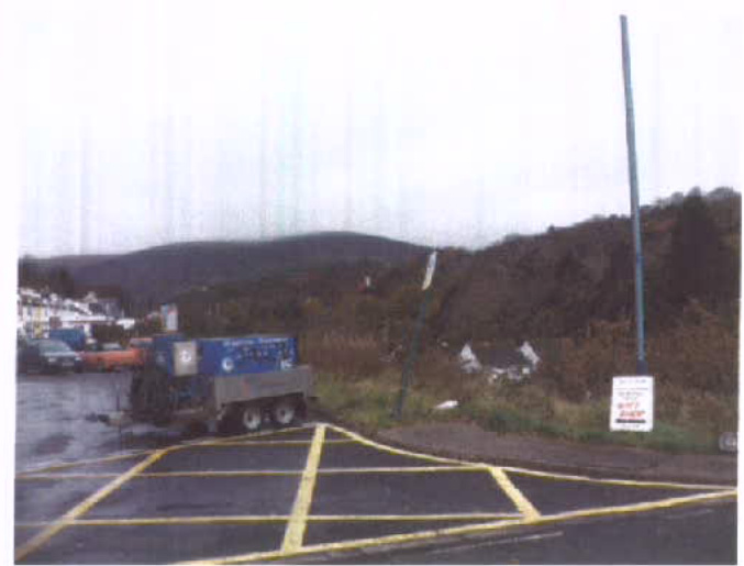 A photograph showing a paved area with yellow hatched road markings and a blue generator on a trailer, with a hillside and buildings in the background.