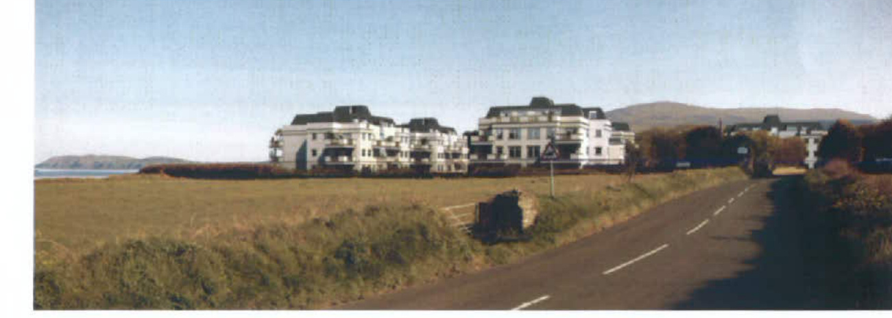 A photograph showing a row of white, multi-story apartment blocks situated along a road with a grassy verge and hills in the background.