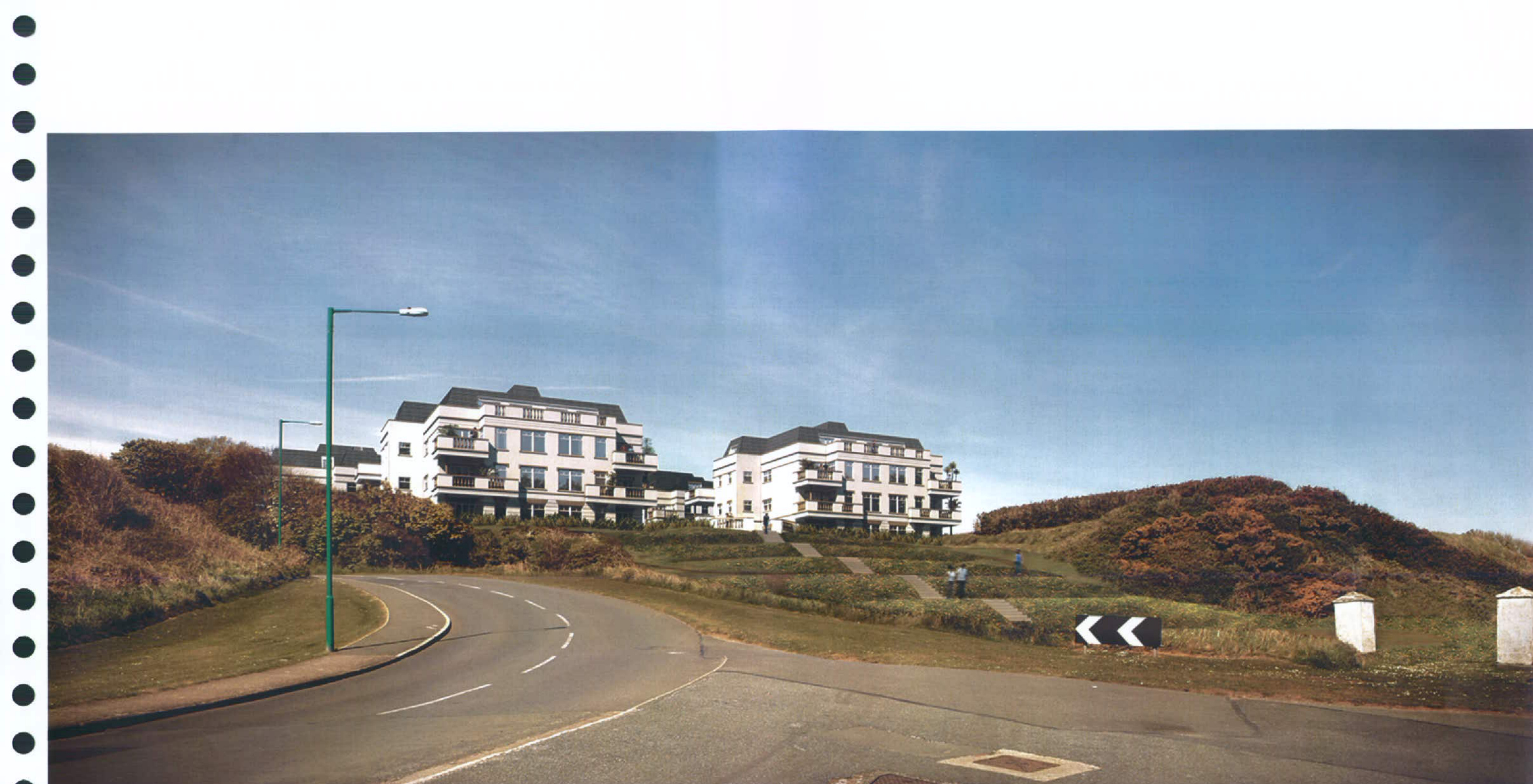 A wide-angle photograph showing a curved road leading past a grassy hillside where several large, white, multi-story apartment blocks are situated under a blue sky.