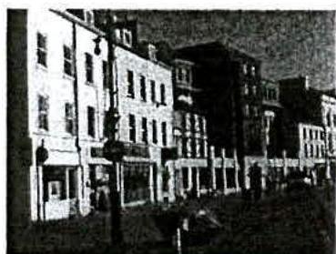 A grainy black and white photograph depicting a street scene with a row of multi-story buildings and pedestrians.
