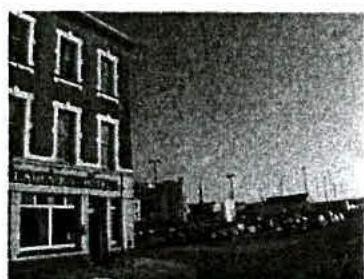A grainy black and white photograph showing the facade of a multi-story building on the left and a street scene with parked cars in the background.