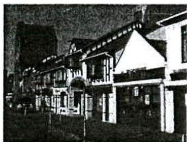 A grainy black and white photograph showing a street scene with a row of terraced buildings and a taller structure in the background.