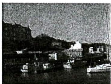 A grainy, low-resolution black and white photograph depicting a harbor or waterfront scene with boats moored in the foreground and buildings on a hillside in the background.