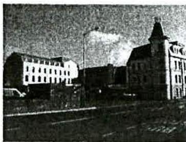 A grainy black and white photograph depicting a street scene with existing buildings, including a prominent building with a tower on the right side.
